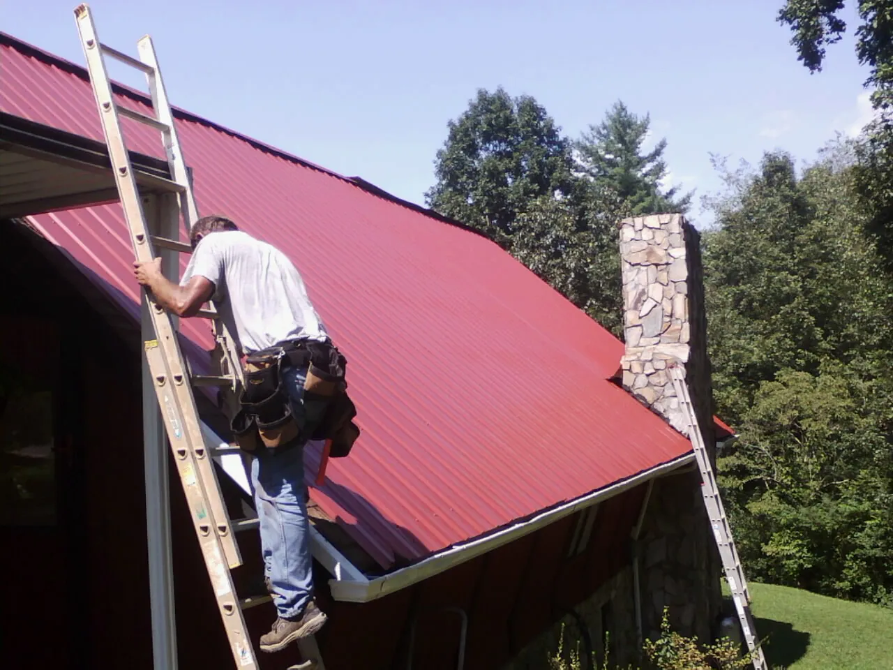 Roofer on a ladder beside a red metal roof and stone chimney — Soffit & Fascia Repair by Roofing Brothers in Portola Valley